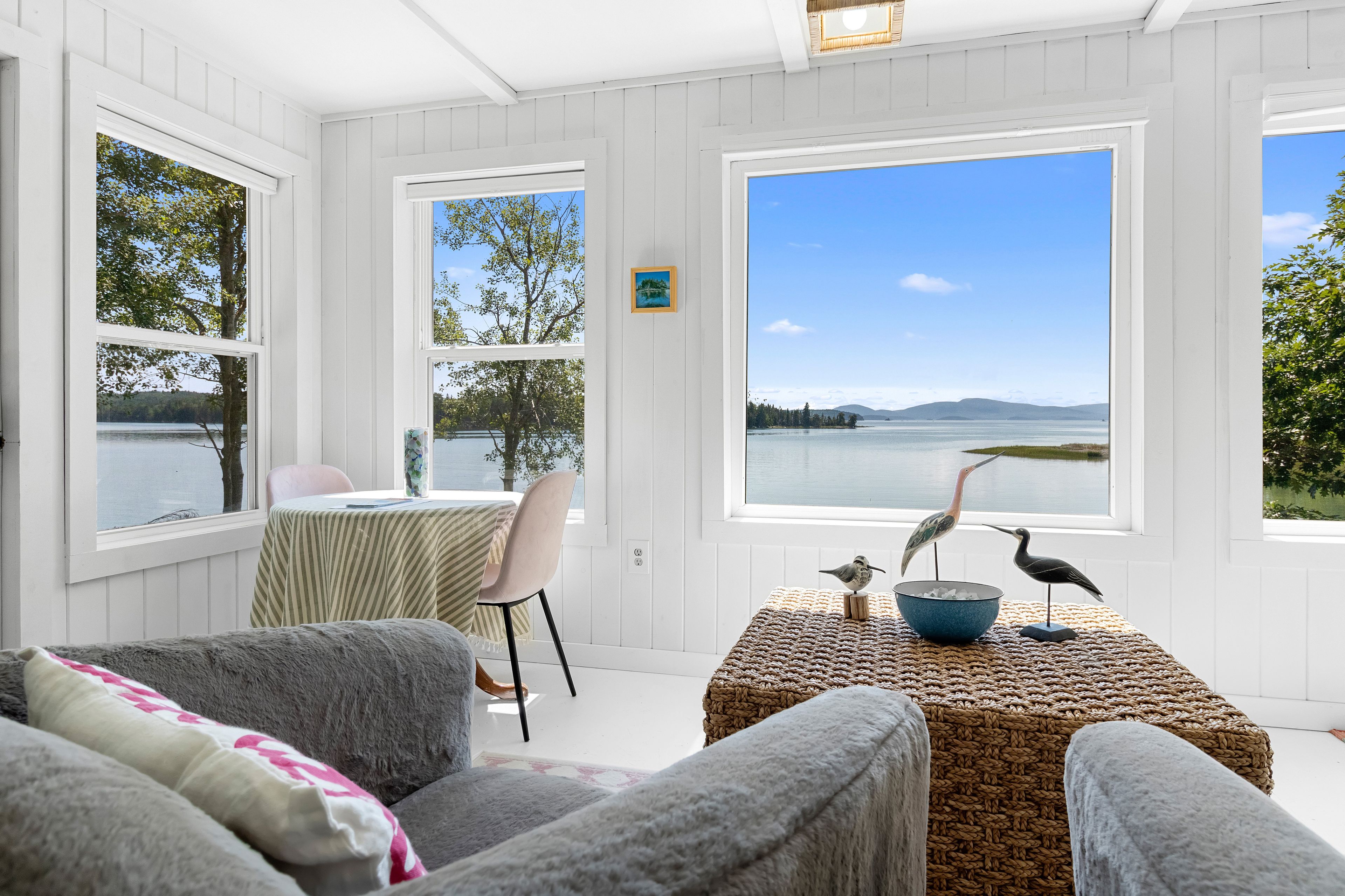 A room with a large window overlooking a lake, a table with a bowl and two bird sculptures, and a chair.