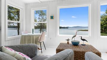 A room with a large window overlooking a lake, a table with a bowl and two bird sculptures, and a chair.