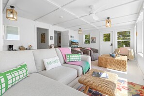 Een ruime woonkamer met een bank, een salontafel en een rieten poef. Er zijn meerdere deuren naar andere kamers, en een televisie is aan de muur bevestigd.