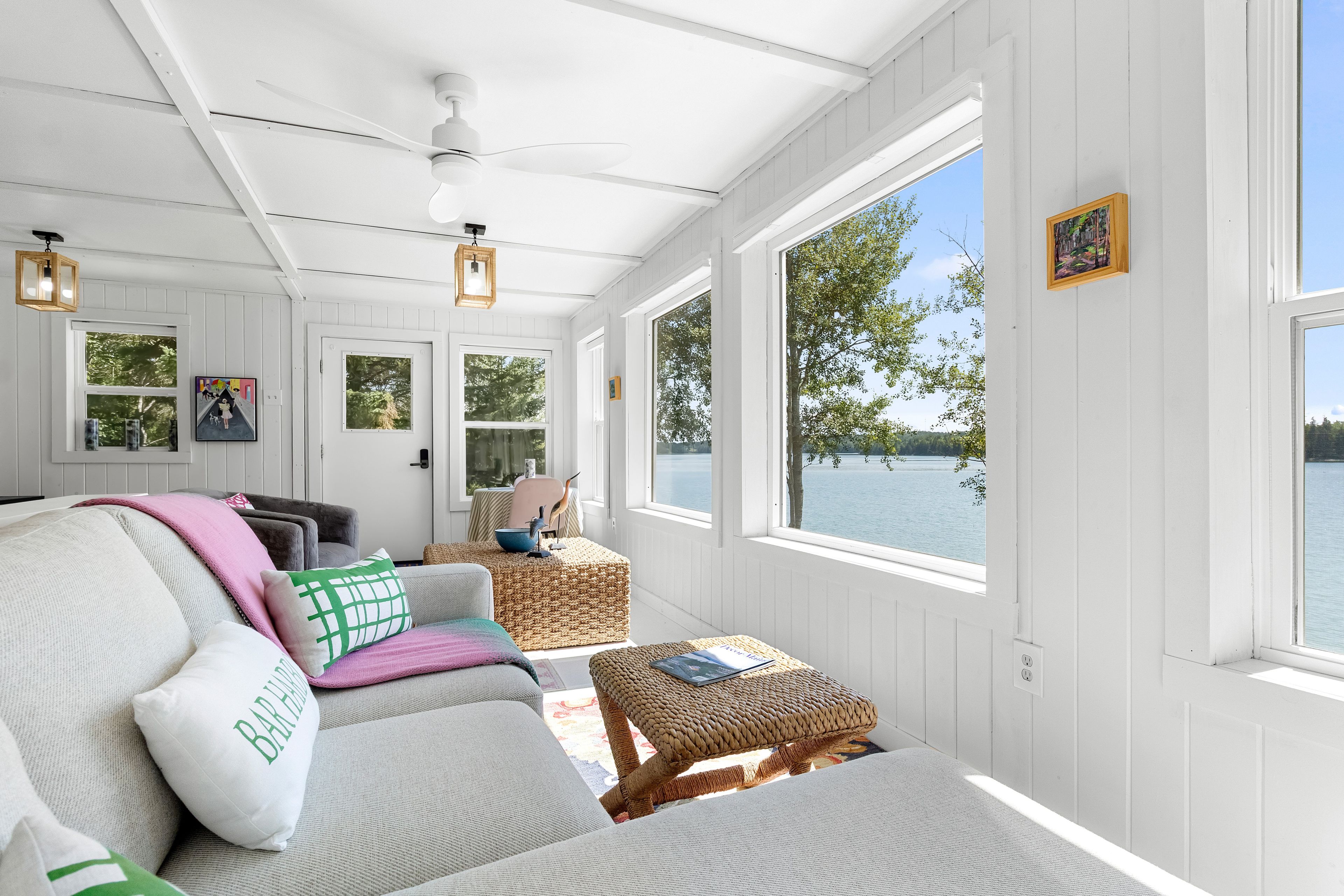 A sunlit room with a view of a lake, featuring a sofa, a wicker ottoman, and a ceiling fan.
