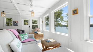 A sunlit room with a view of a lake, featuring a sofa, a wicker ottoman, and a ceiling fan.