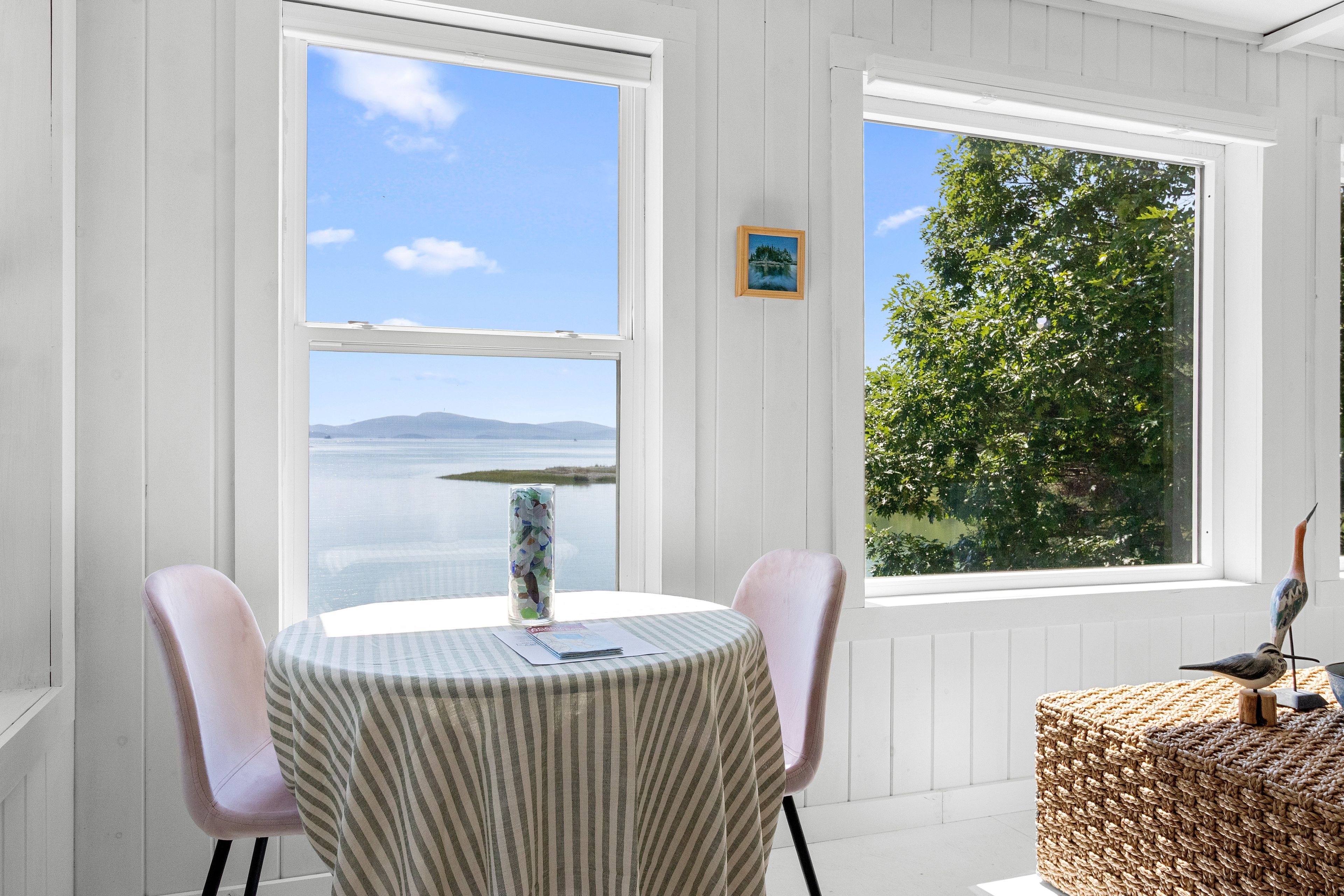 A room with a round table, two chairs, a window with a view of a body of water and mountains, and a framed picture on the wall.