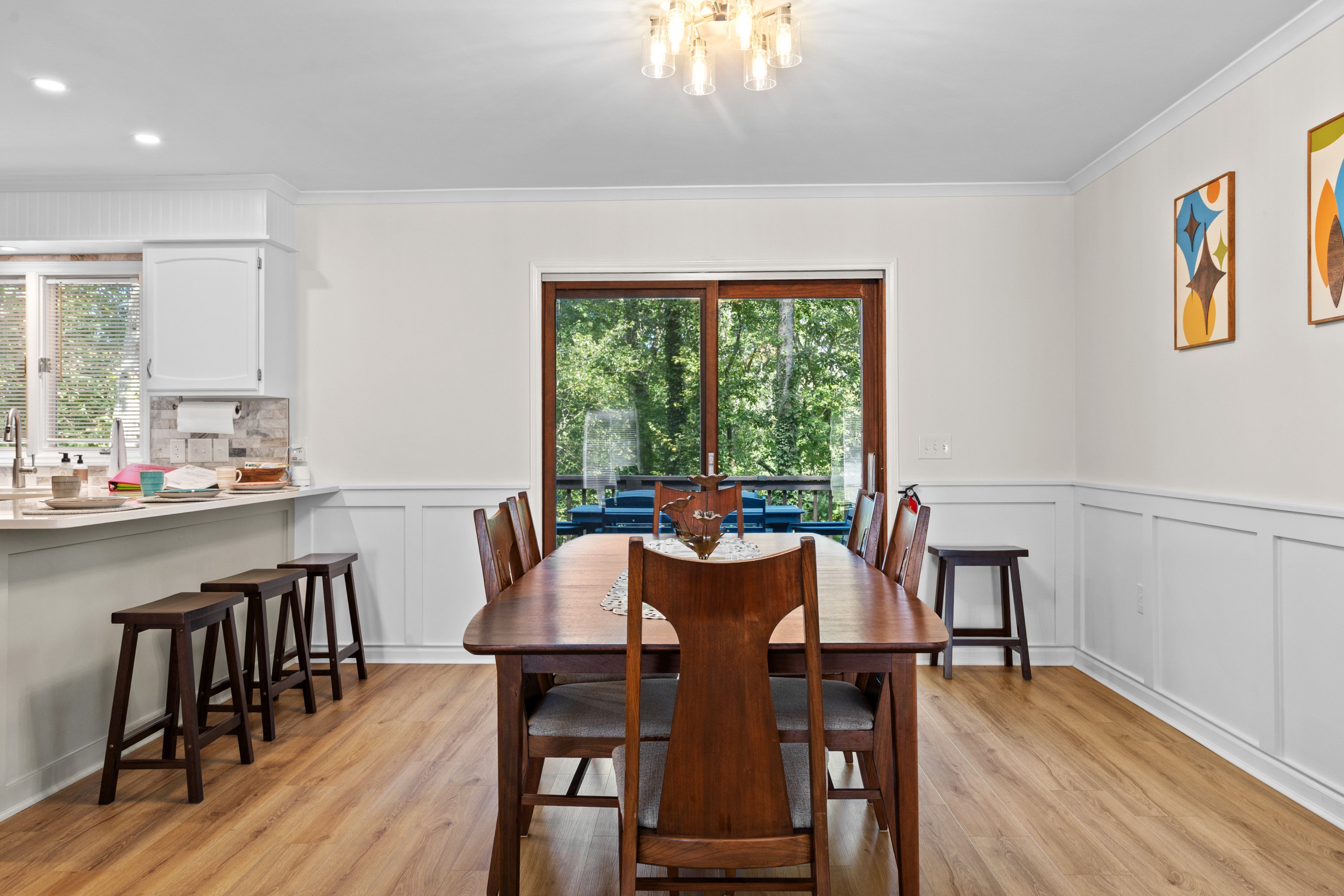 A dining room with a wooden table and chairs, a kitchen area with white cabinetry, and a view of the outdoors through a large window.