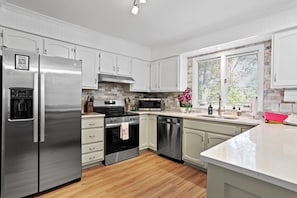 A modern kitchen with white cabinetry, stainless steel appliances, a stone backsplash, and a large window with blinds. - Twin Pines of Hendersonville (Hendersonville)
