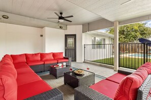 A covered patio with a red sectional sofa, two side tables, and a ceiling fan. - Private Pool & Loungers | Theater, Dry Sauna & Games (San Antonio)