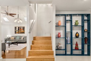 A modern living room with a sofa, coffee table, and artwork. A wooden staircase leads to a hallway with a blue shelving unit displaying various decorative items. - Private Pool & Loungers | Theater, Dry Sauna & Games (San Antonio)