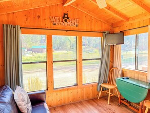 Interior de una cabaña con techo de madera, un sofá, un escritorio y amplios ventanales que ofrecen vistas al exterior.