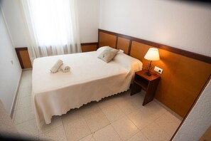 Ein ordentlich bezogenes Bett mit Kissen und zwei gerollten Handtüchern, ein Nachttisch mit Lampe sowie ein Fenster mit durchsichtigen Vorhängen.