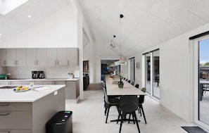 A modern kitchen with a central island, a dining area with a long table, and a view through glass doors to an outdoor patio. - Beautiful home in Idestrup with sauna (Idestrup)
