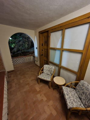 Una habitación con puerta de madera marcada con el número '38', una silla estampada, una pequeña mesa redonda y vista a árboles a través de un ventanal de arco.