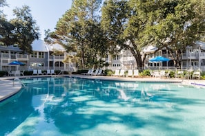 A swimming pool with lounge chairs and umbrellas, surrounded by trees and a building with balconies. - Ocean Palms Luxury Villa-Daily Free Golf+Tennis (Hilton Head Island)
