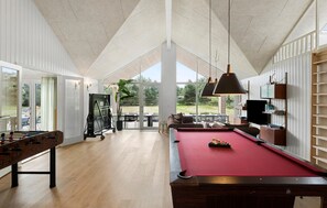 Un grand espace intérieur comprenant une table de billard, un baby-foot et un coin repas offrant une vue sur l’extérieur.