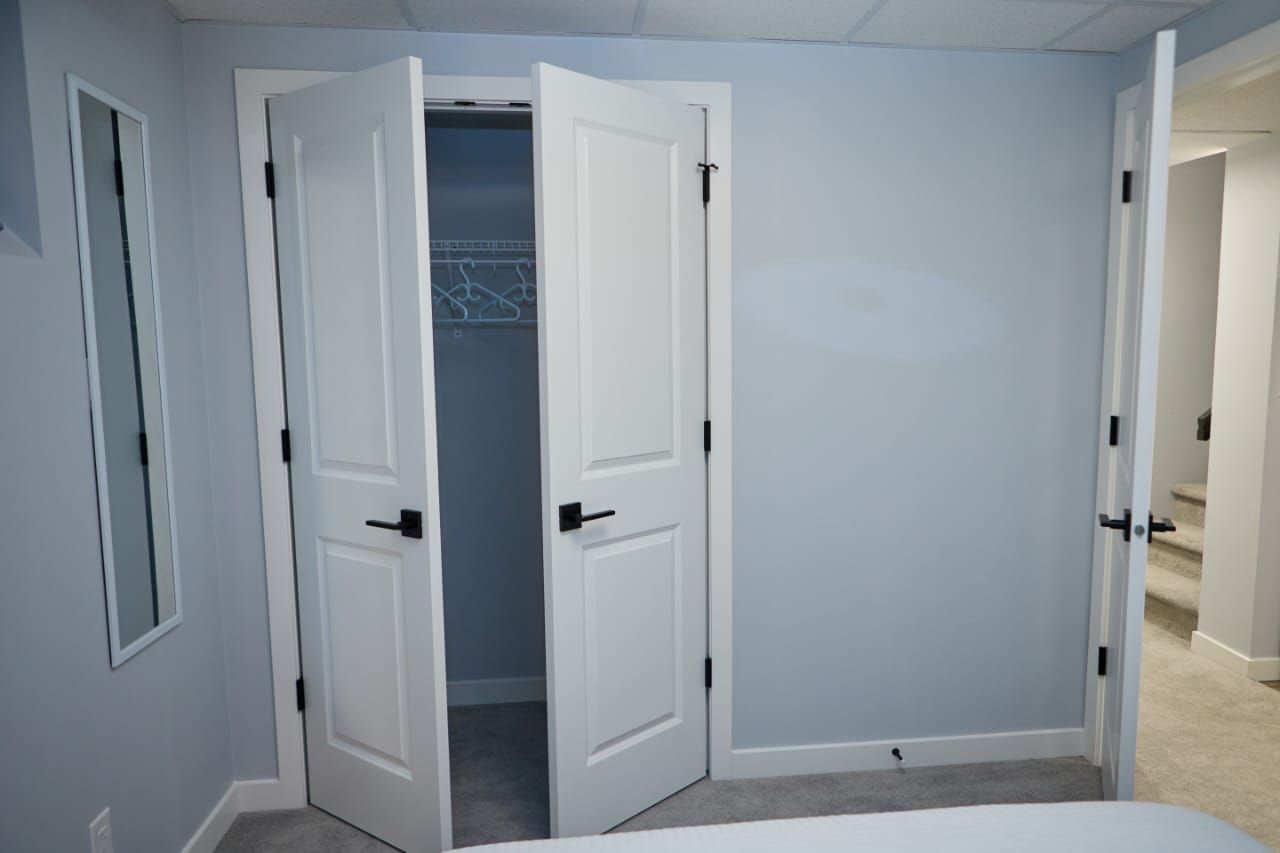 A room with two white doors, one open revealing a closet with hanging clothes, and a carpeted floor.