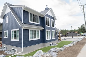 A two-story residential building with a gable roof, large windows, and a front yard with rocks and grass. - B.C. Basecamp | Mountain Escape • Sleeps 15 (East Kootenay)
