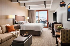 Ein Hotelzimmer mit Bett, Sofa, Schreibtisch, Fernseher und Blick auf eine verschneite Landschaft.