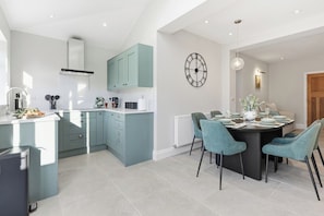 A modern kitchen with teal cabinetry, a dining area with a round table, and a wall clock. - Luxury 4-Bed Contractor/Family House Sleeps 7 (Cheshire West and Chester)