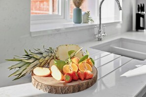 Een keukentafel met een fruitstuk op een houten plank, een spoelbak en een raam met uitzicht.