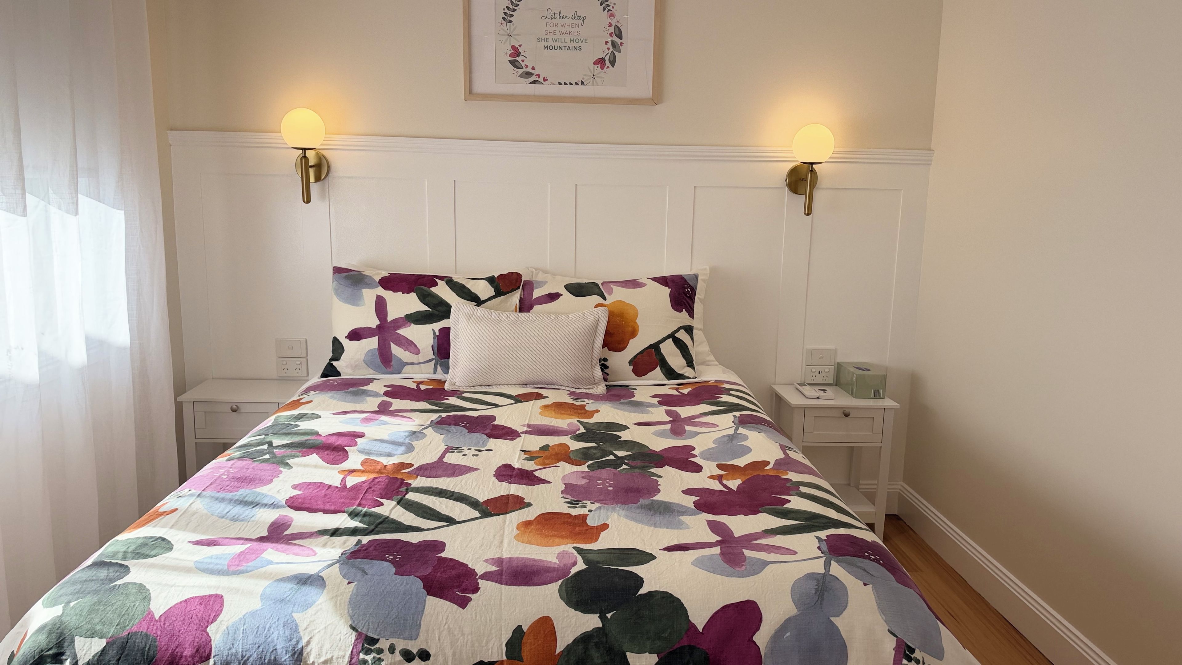A bed with a floral bedspread, two bedside lamps, a framed quote on the wall, and a nightstand with a box.