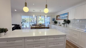 A modern kitchen with white cabinetry, a central island, pendant lighting, and a dining area with a view to the outdoors. - Holiday House Southport near Surfers Paradise. (Southport)