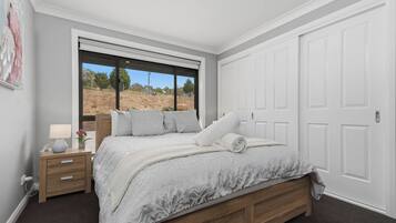 Ein Schlafzimmer mit großem Fenster, einem Bett mit weißer Bettdecke, einem Nachttisch mit Lampe und einer Tür.