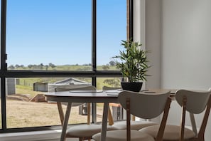 Een eethoek met een tafel en stoelen, een potplant en uitzicht op een landelijk landschap door grote ramen.