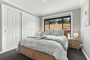 Een slaapkamer met een groot bed, een houten hoofdbord, een nachtkastje met een lamp en uitzicht op een buitenruimte door een raam.
