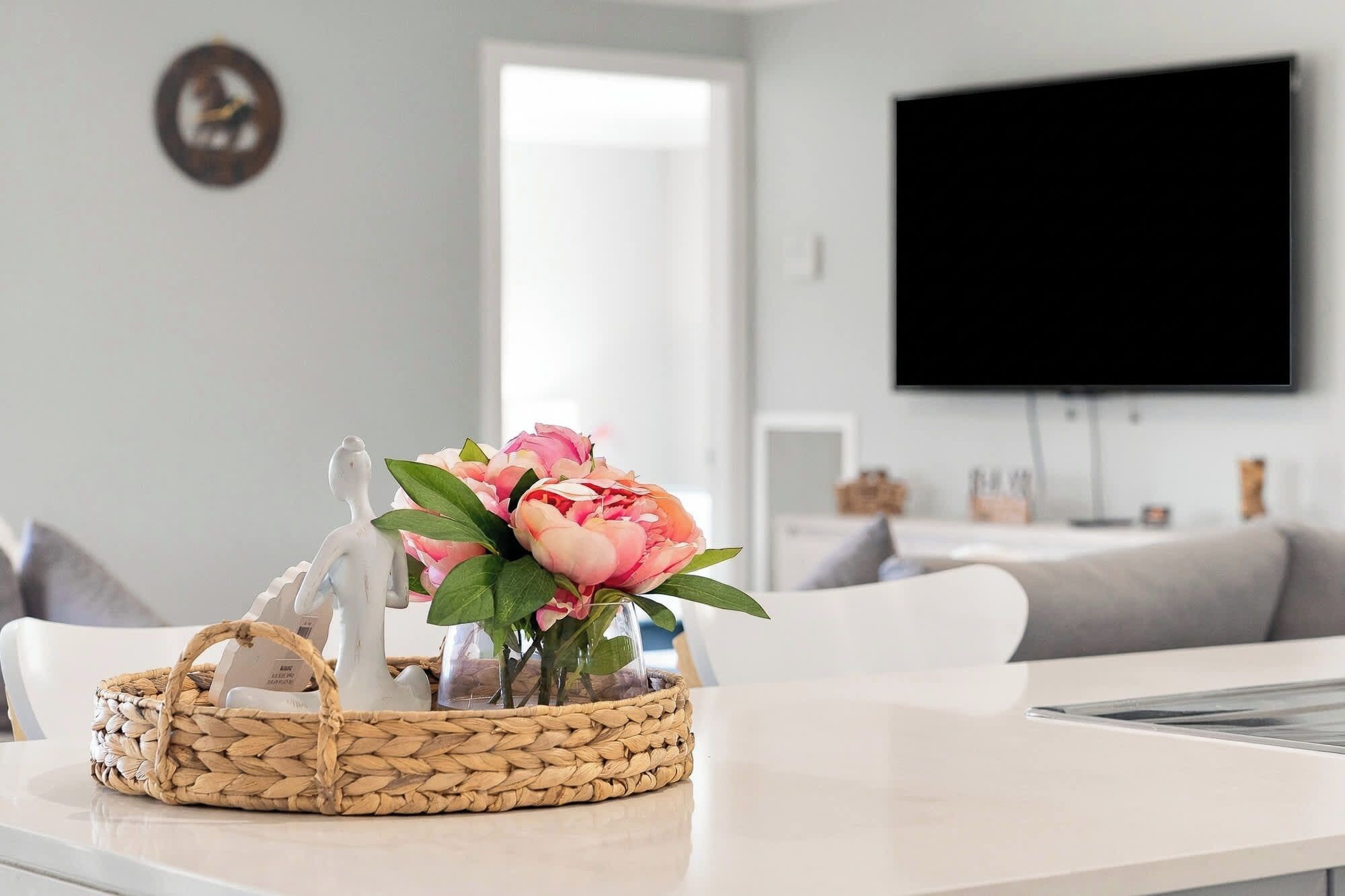 Un soggiorno con un divano bianco, una TV a schermo piatto montata a parete e un cesto di vimini con un vaso di fiori su un tavolo.