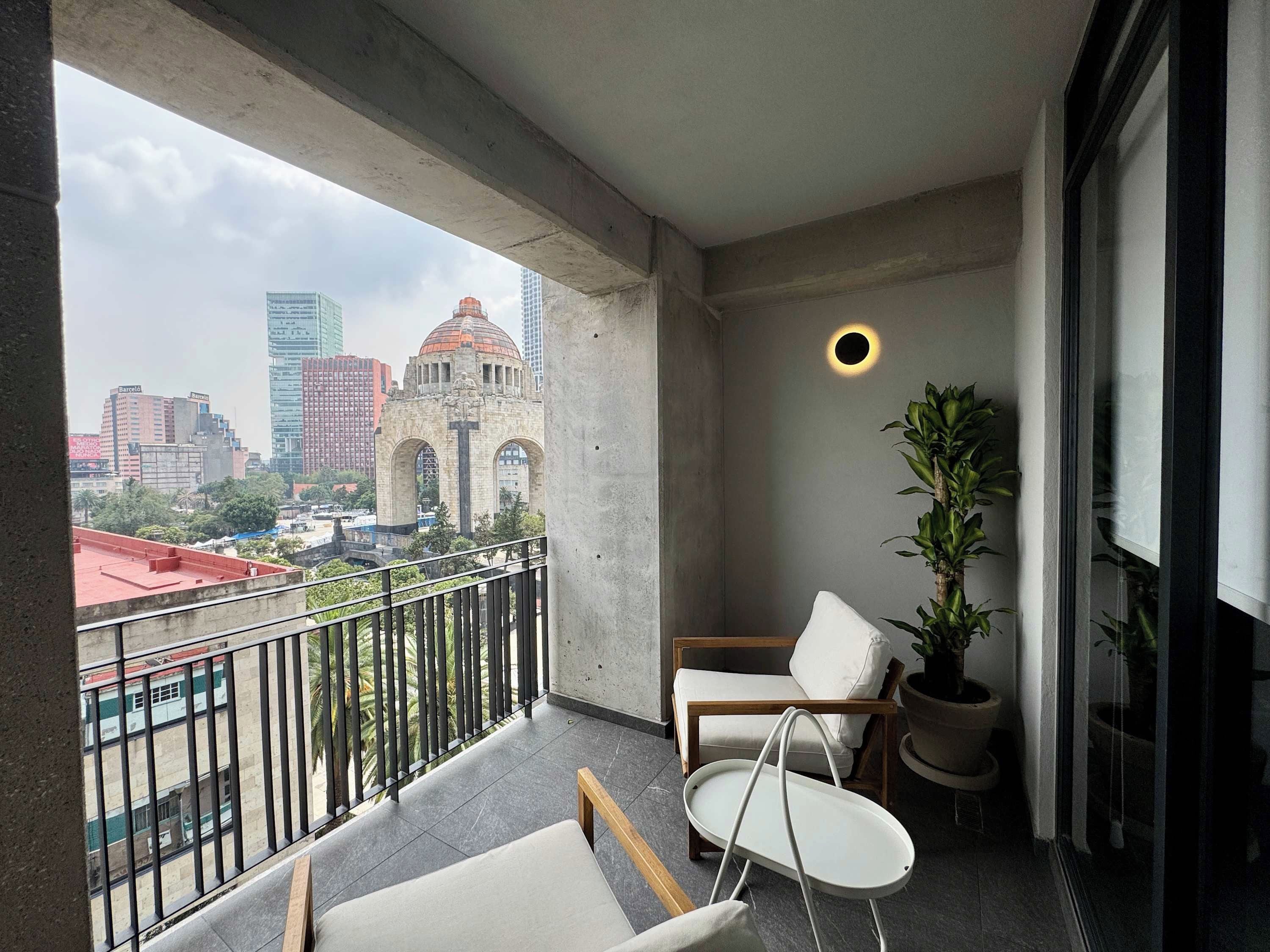 Un balcón con vista a un paisaje urbano, incluyendo un edificio destacado con cúpula. Hay una planta en maceta y una silla blanca.