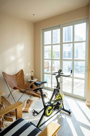 A room with a stationary bike, a brown leather chair, a wooden bench, and a large window with a view of buildings. - w* | Upscale 3BR in Prime Bocagrande (Cartagena de Indias)