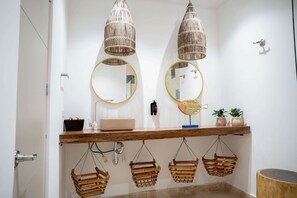 Um banheiro com duas luminárias de vime suspensas, um espelho redondo com moldura dourada, uma touca de madeira com pia e três cestos de vime pendurados.