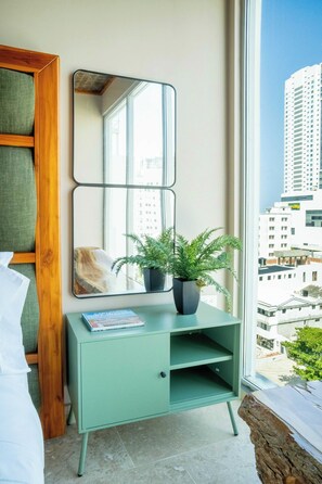 Ein Zimmer mit einem grünen Konsolentisch, einem Spiegel, einer Topfpflanze und Blick auf eine Stadtkulisse durch ein großes Fenster.