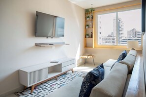 A modern living room with a flat-screen TV, a sofa with patterned cushions, a small side table, and a window with a city view. - w* | Luxury 3BR in Prime Bocagrande (Cartagena de Indias)