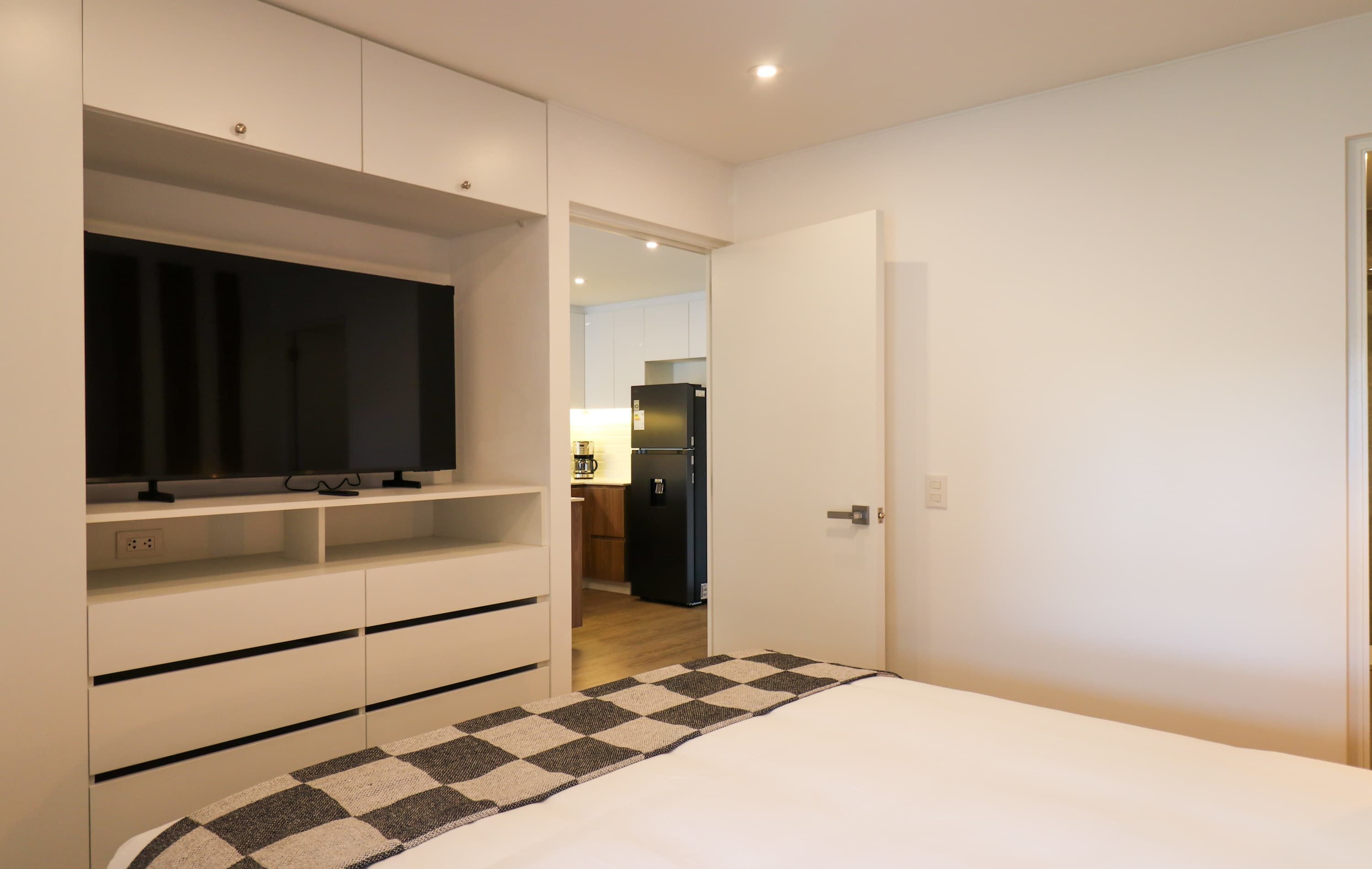 A bedroom with a bed, a television on a stand, and a kitchen in the background.