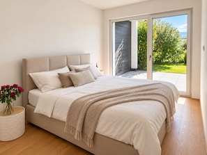 A bedroom with a bed, a bedside table with a vase of flowers, and a view of a garden through a sliding door. - Premium garden apartment on the Kreuzbergl near Lake Wörthersee, the University, and Lakeside (Klagenfurt am Wörthersee)