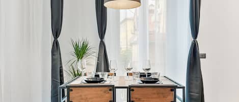 A dining area with a table set for two, featuring a modern pendant light, a potted plant, and a view of the city through large windows.
