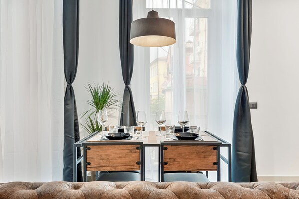 Ein Essbereich mit Tisch für zwei Personen, ausgestattet mit einer modernen Pendelleuchte, einem Topf mit Pflanze und Blick auf die Stadt durch große Fenster.