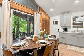 A dining area with a wooden table set for four, a kitchen with grey cabinets, and a view of greenery through the windows. - Rustic Lakeside Cabin | Dock + Kayaks | Locke Lake (Barnstead)