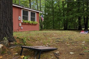 A red cabin with a flag, surrounded by trees and a picnic table. - Rustic Lakeside Cabin | Dock + Kayaks | Locke Lake (Barnstead)