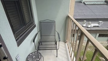 A balcony with a metal chair and a small table, a window with blinds, and a building in the background.