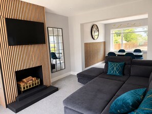 A modern living room with a grey sofa, a fireplace with a wooden panel design, a large mirror, and a dining area with blue chairs. - Modern 2-bed House With Garden Quiet Street, Easy London & Watford Access (Watford)