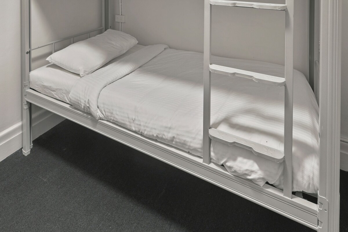 Una cama litera con escalera, ropa de cama blanca y un enchufe en la pared.