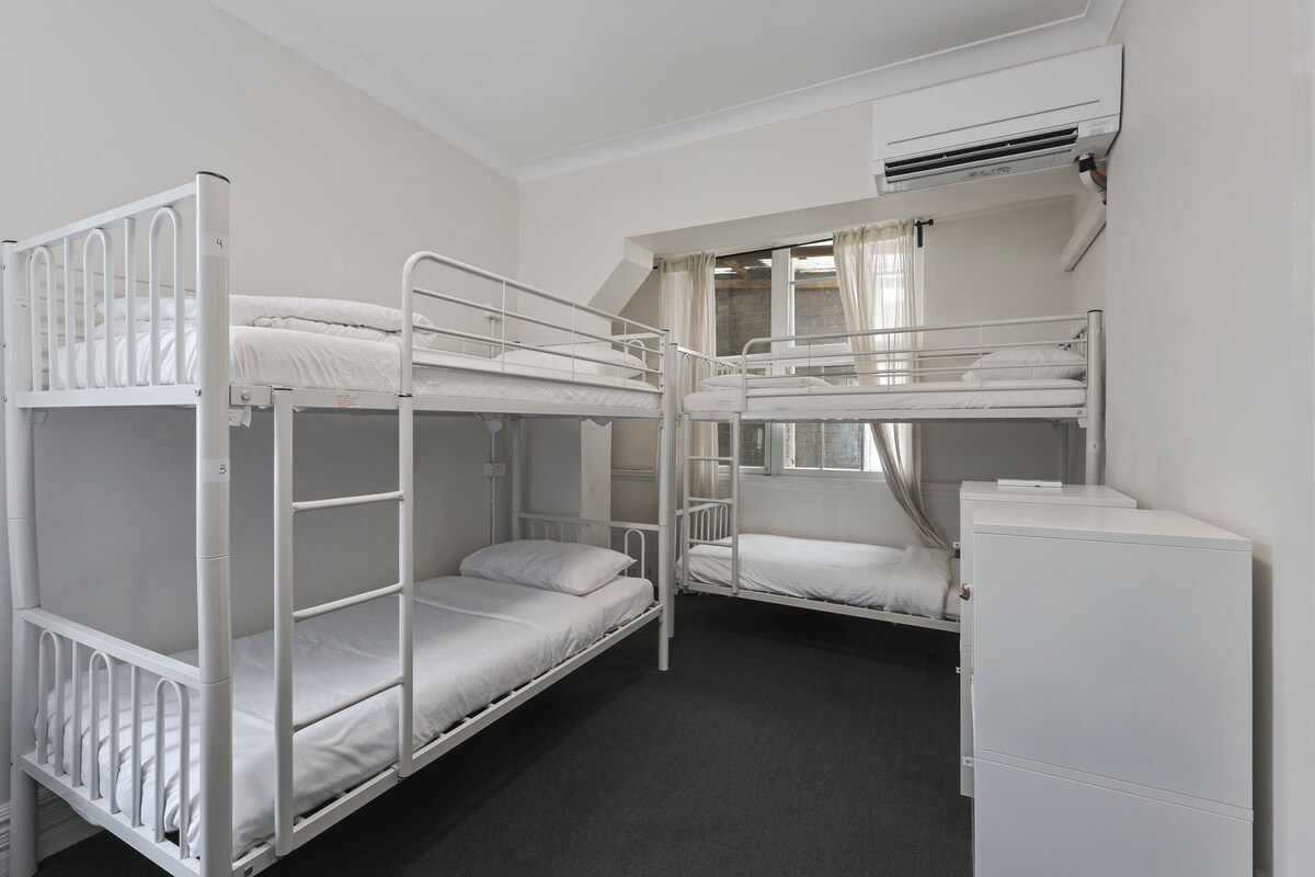 Un dormitorio con literas, tres literas, un mueble blanco pequeño y un equipo de aire acondicionado en la pared.