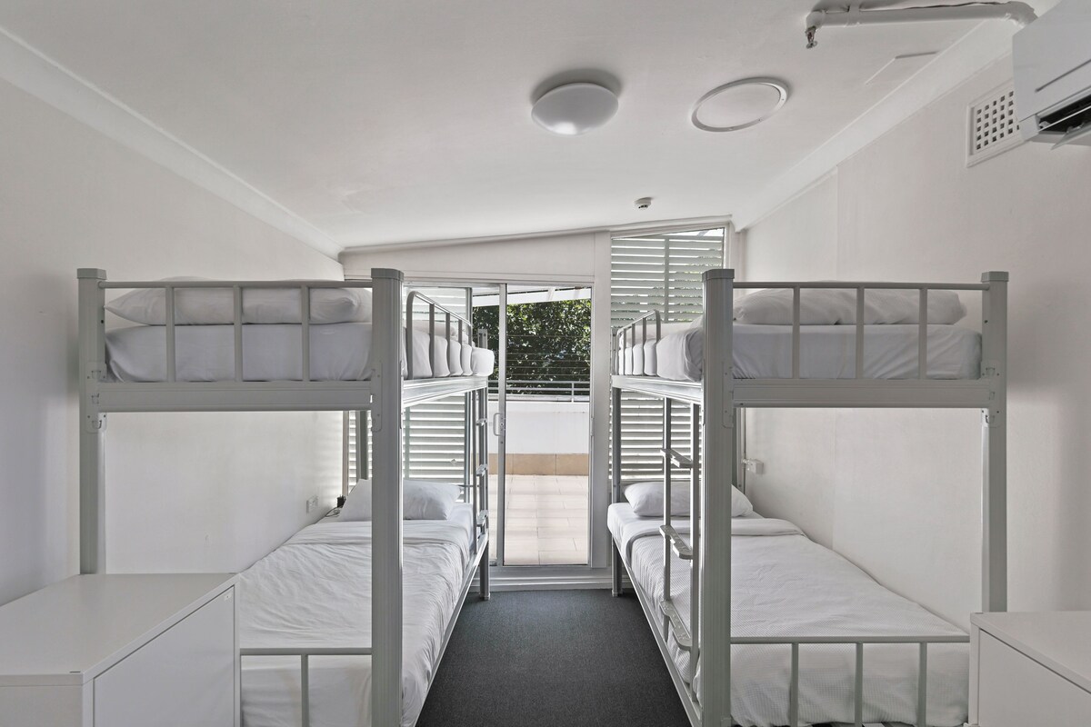 Dormitorio con dos literas, una lámpara en el techo y una ventana con persianas.