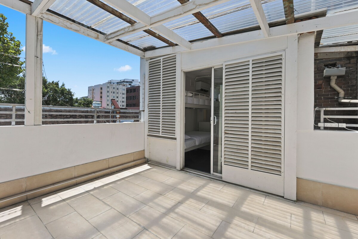 Un balcón con cerramiento de vidrio, una puerta corredera y vistas a edificios al fondo.
