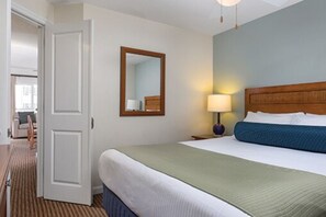 Een hotelkamer met een bed, nachtkastje, lamp, spiegel en een deur die naar een andere kamer leidt.