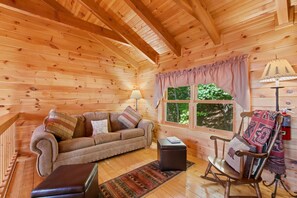 Ein gemütliches Blockhauss-Interieur mit Holzbalkendecke, einer Couch, einem Schaukelstuhl, einem kleinen Tisch und einem Fenster mit Blick auf Bäume.