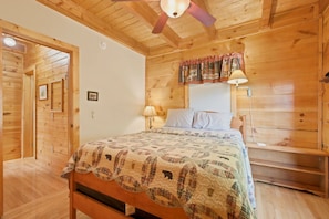 Ein Schlafzimmer einer Holzhütte mit einem Bett, einem Nachttisch, einer Lampe und einem Fenster mit Vorhängen.