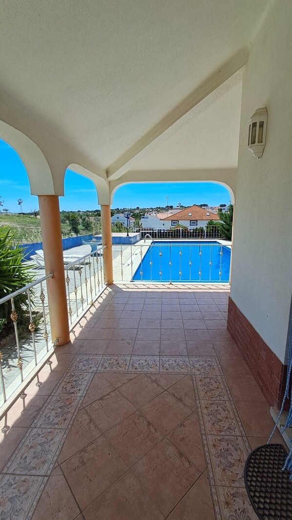A covered balcony with a view of a swimming pool and houses in the distance.