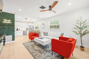A modern living room with a white sofa, red armchairs, and a coffee table. There is a ceiling fan, a green wall, and a kitchen in the background. - Coastal Getaway - 13 Min to Beach - Bike Trail (Belleair)