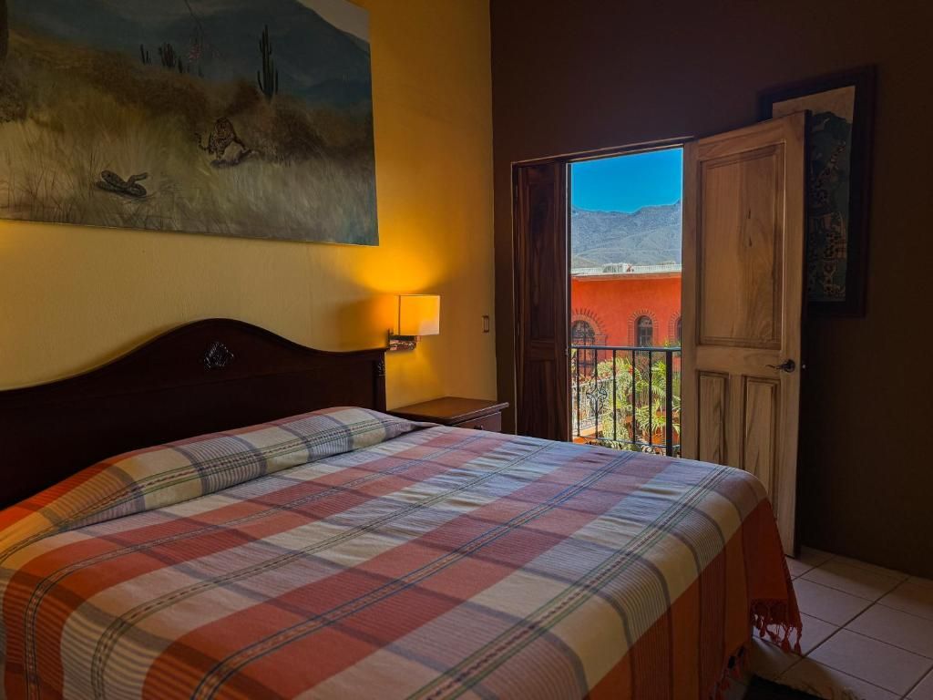 Un dormitorio con cama, mesita de noche, un cuadro, una puerta que da a un balcón y vista a un edificio.